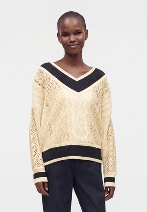 Smilende person iført en cremefarvet strikket V-hals sweater med sort kant og mørke bukser, stående mod en ensfarvet baggrund.