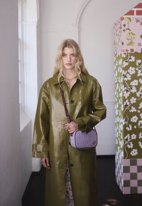 Lesklý koženkový kabát olivově zelené barvy s uvolněným střihem a velkými knoflíky. Doplněný o malou fialově pruhovanou kabelku přes rameno.