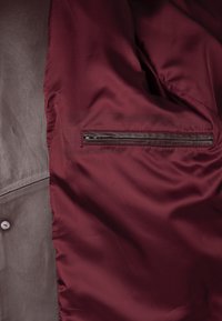 Bordeauxrotes Innenfutter einer Jacke mit glatter Textur und einer Reißverschlusstasche. Der Außenstoff wirkt grau und leicht strukturiert.