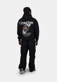 Černá mikina s kapucí s bílým nápisem "Crystal Paris" a designem hodinek na zadní straně, kombinovaná s černými cargo kalhotami a vícetrojbarevnými teniskami.