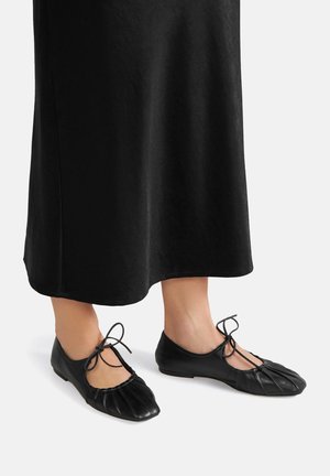 Černé kožené baletní baleríny s skládacím vzorem, mají kulatou špičku a tenké tkaničky pro bezpečné uchycení, v kombinaci s dlouhou černou sukní.