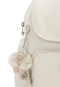 Mochila beige con cierre de cremallera, que cuenta con un bolsillo frontal y un llavero de mono de peluche como accesorio. Tejido suave y detalles de metal.
