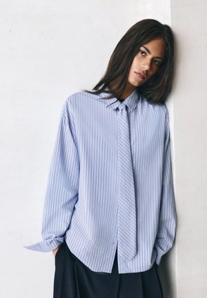 Tânăr cu păr lung și închis la culoare se sprijină de un perete alb, purtând o cămașă largă albastru deschis cu dungi subțiri și o cravată asortată, cu mâinile în buzunare.