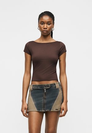 Kvinde med fletninger iført brun tætsiddende kortærmet top og tofarvet denim mininederdel med lynlås foran, står mod en ensfarvet baggrund.