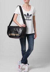 Person som bär en vit Adidas t-shirt, blåjeans, vita Adidas sneakers med röda ränder, och bär en svart Adidas axelväska med guld-logotyp.