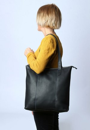 Schwarze Ledertasche mit glatter Oberfläche, ausgestattet mit blauen Nähakzenten und einem Reißverschluss, die lässig über einer Schulter getragen wird.