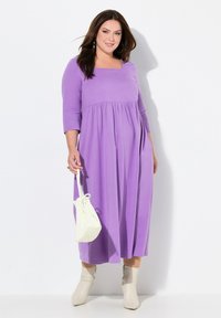 Violetti, pitkä, löyhästi istuva mekko, jossa on kolme neljännesosaa hihat ja neliön muotoinen kaula-aukko. Yhdistetty valkoiseen käsilaukkuun ja beigeihin, vetoketjullisiin nilkkureihin.