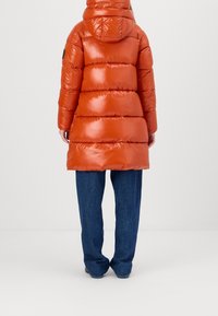 Long puffer coat in shiny orange with a hood, featuring horizontal quilted sections. Paired with loose blue denim pants.