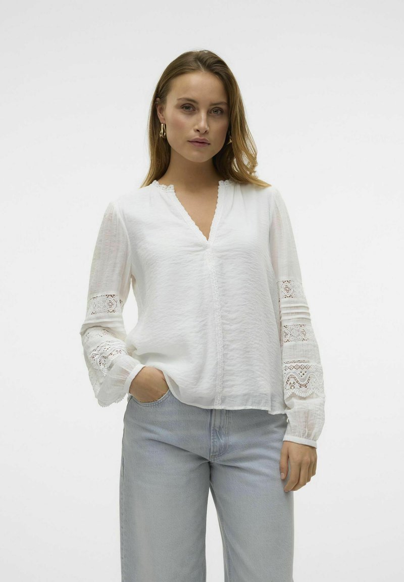 Blusa bianca con scollatura a V, tessuto texture e dettagli in pizzo sulle maniche lunghe. Abbinata a jeans a gamba larga blu chiaro.