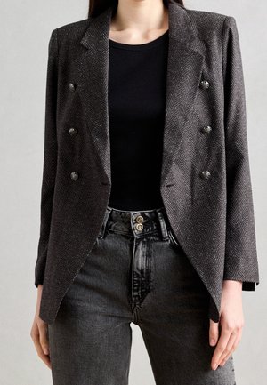 Kvinde iført en mørkegrå fiskebensmønstret dobbeltradet blazer over en sort top og højtsiddende falmede sorte jeans.