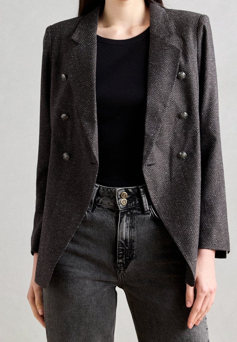 Femme portant un blazer croisé en chevrons gris foncé sur un top noir et un jean noir délavé taille haute.