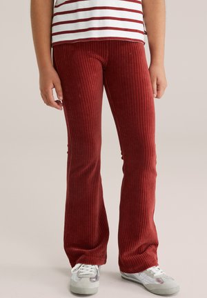 Bambino con pantaloni in velluto a coste rossi, sneakers bianche e una maglietta bianca a righe orizzontali rosse, in piedi davanti a uno sfondo semplice.