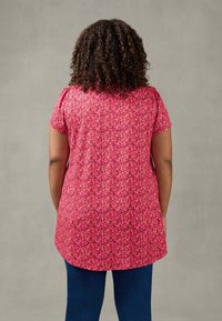 Short-sleeved tunic in vibrant pink floral fabric, with a relaxed fit and curved hem. Paired with dark jeans, showcasing a textured pattern.