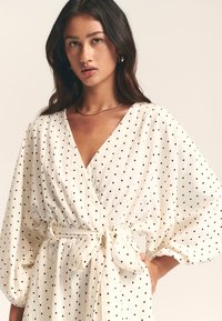 Robe portefeuille blanche à pois noirs, manches longues bouffantes, ceinture à nouer à la taille et décolleté en V profond. Texture de tissu lisse.