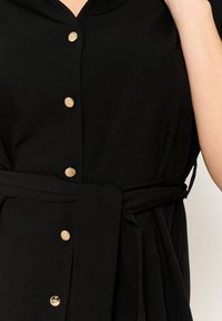 Czarna sukienka zapinana na guziki, z dekoltem w serek, krótkimi rękawami, złotymi guzikami i materiałowym paskiem przewiązanym w talii. Gładka faktura, brak widocznych wzorów.