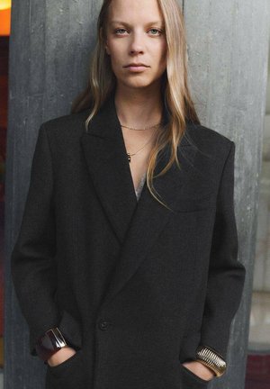 Blazer de lana negra con hombros estructurados, cierre de un solo botón y dos bolsillos. El modelo lleva pulseras doradas y oscuras. Fondo texturizado.