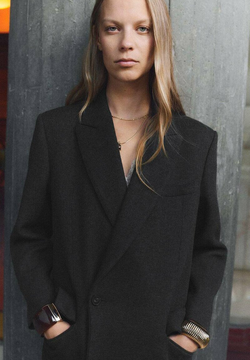 Sacou negru din lână cu umeri structurați, închidere cu un singur nasture și buzunare duble. Modelul poartă brățări aurii și închise la culoare. Fundal texturat.
