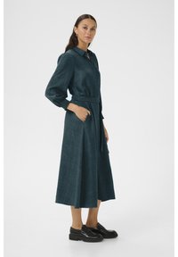 Teal, pitkähihainen, napitettava mekko, jossa on vyötäröllä vyö, sivutaskut ja teksturoitu kangas. Mallilla on päällä mustat korokepohjaiset kengät.