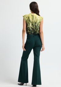 Vrouw die staat, gekleed in een mouwloze groen-geel top met bladdessins en donkergroene wijde broek met zwarte hakken.
