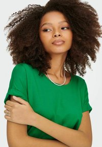 Nő dús göndör hajjal, zöld ingben, gyöngy nyaklánccal és ezüst gyűrűvel, oldalra néz, karját keresztbe téve.