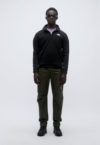 Μαύρο φλις πλεκτό με φερμουάρ και λογότυπο, παντελόνι σε ελιά πράσινο, και μαύρα παπούτσια. Το μοντέλο φορά γυαλιά ηλίου· ουδέτερο φόντο.