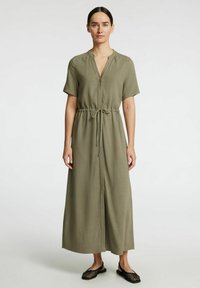 Robe maxi vert olive avec manches courtes, décolleté en V, taille à cordon et coupe décontractée. Portée avec des sandales noires à bout ouvert en maille.