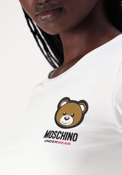 Biele tričko s krátkym rukávom, na hrudi s grafikou hnedého medveďa a nápisom "MOSCHINO UNDERBEAR" v čiernej a červenej farbe. Hladká textúra látky.