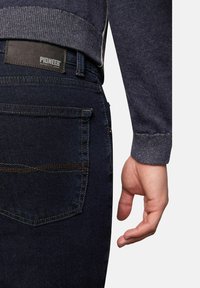 Donkerblauwe denimjeans met een leren label "Pioneer" aan de achterkant. De trui is grijs met een geribde manchette. Hand in de buurt gepositioneerd.