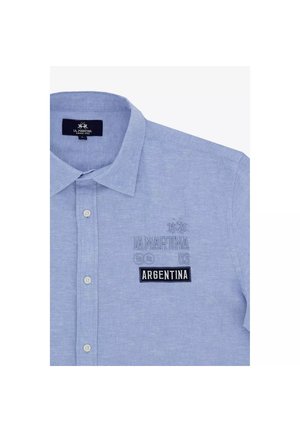 Lichtblauw overhemd met knopen en kraag, geborduurd met "LA MARTINA 09" en een zwarte patch met de tekst "ARGENTINA" op de linkerborst.