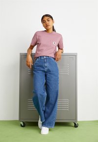 Maglietta rosa a maniche corte con taschino e logo, abbinata a jeans blu a gamba larga. Lo sfondo semplice presenta un armadietto metallico grigio.