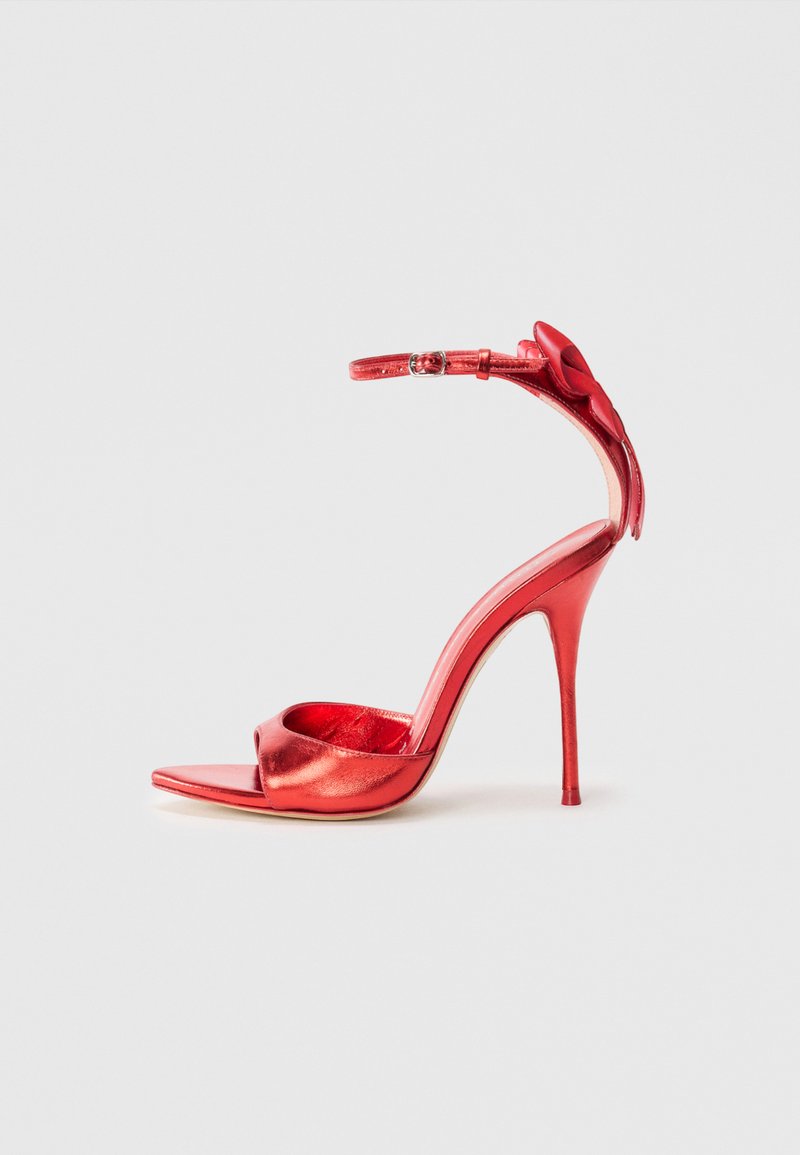 Punaiset metalliset korkokengät, joissa on avonainen varvas, säädettävä nilkkahihna ja koristeellinen nauha kantapäässä. Pintamateriaali on sileää.