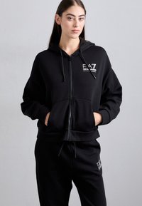 Žena u crnoj majici s patentnim zatvaračem i odgovarajućim trenirkom sa bijelim EA7 Emporio Armani logotipima, stoji naslonjena na jednoličnu svijetlosivu pozadinu.