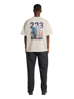 Mężczyzna zwrócony tyłem, ubrany w biały oversize T-shirt z dużym napisem „223” i grafiką miasta i gór na plecach, czarne spodnie i czarne buty.