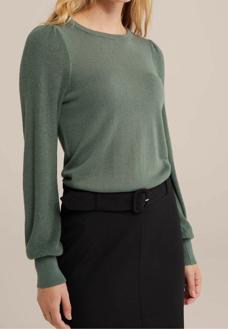 Grön stickad tröja med långa puffärmar, rund halsringning och ribbade muddar, parat med ett svart bälte och kjol, vilket skapar en strömlinjeformad silhuett.