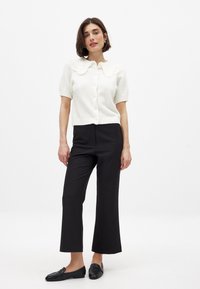 Bela kratka kardigan s širokim teksturiranim ovratnikom, s gumbom za zatvaranje, uparen z črnimi širokimi hlačami in črnimi loaferji.