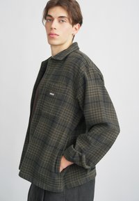 Zeleno-črna karirana flanelasta jakna, z zadrgo, ovratnikom, stranskimi žepi, vezanim logotipom na prsih, sproščen kroj in teksturirana tkanina.