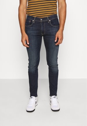 Homme portant un jean slim bleu foncé, une chemise rayée marron et jaune, et des baskets blanches Nike, debout sur un sol blanc.