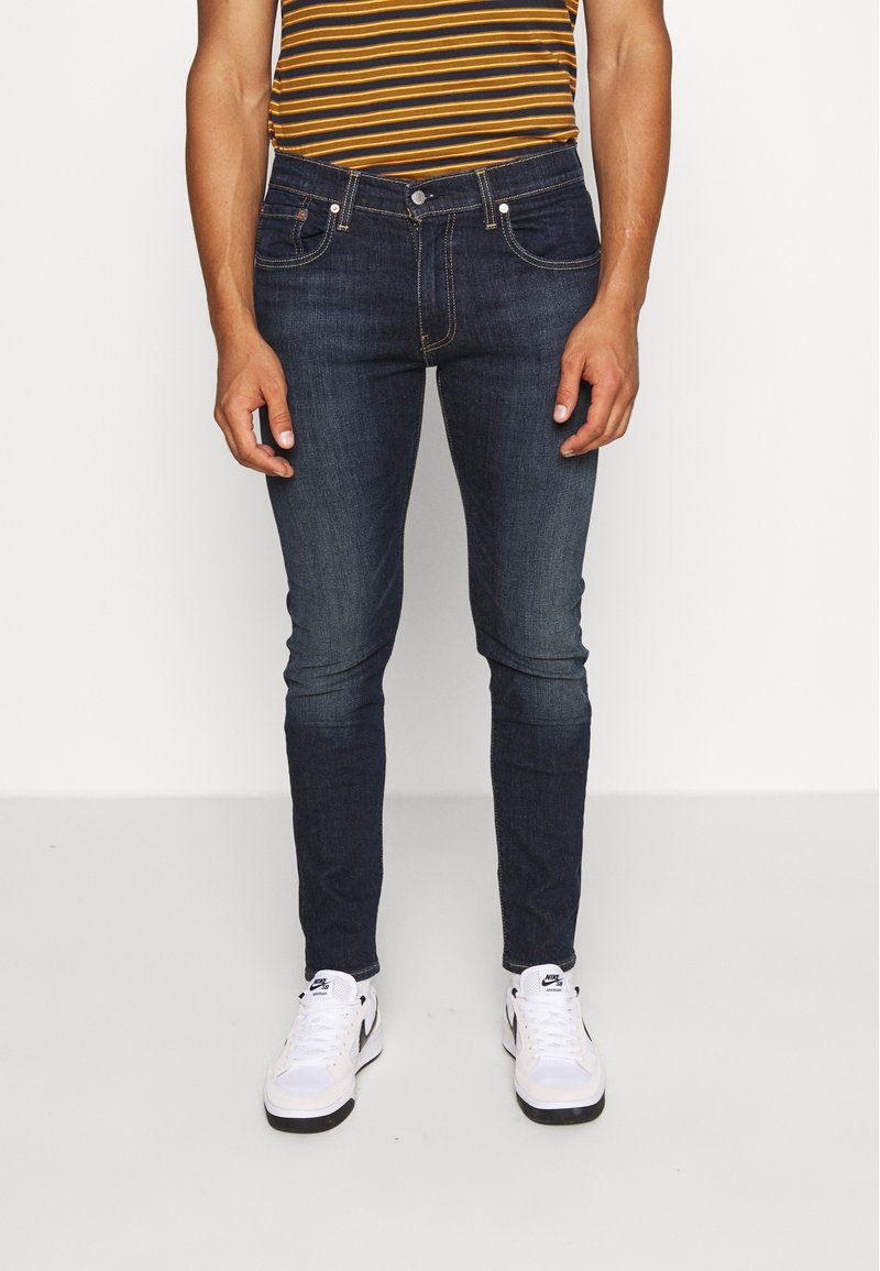 Homme portant un jean slim bleu foncé, une chemise rayée marron et jaune, et des baskets blanches Nike, debout sur un sol blanc.