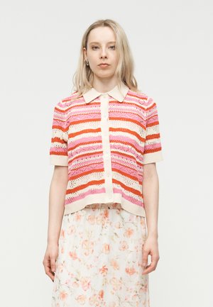 Giovane donna con capelli biondi lunghi che indossa un cardigan a maglia a maniche corte a righe e una gonna bianca floreale, in piedi davanti a uno sfondo semplice.