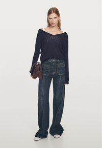 Frau trägt dunkle Schlagjeans, navyfarbigen schulterfreien Pullover, weiße Schuhe und eine braune Handtasche vor hellem einfarbigem Hintergrund.