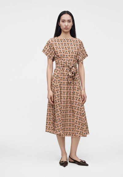 Femme debout portant une robe mi-longue à motifs avec manches courtes et ceinture à la taille, associée à des chaussures slingback noires pointues.
