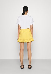 Geel oogjesrok met een gerimpelde zoom, elastische tailleband. Gecombineerd met een wit t-shirt en zwarte sandalen. Achteraanzicht.