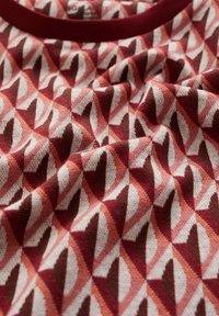 En bomull t-shirt med ett geometriskt mönster av röda, rosa och vita former. Ribbad krage i mörkröd färg. Mjuk textur, som visar vikta tyger.