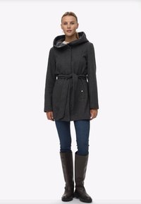 Sieviete stāv tērpusies tumši pelēkā jostā saistītā kapucē, zilos džinsos un tumši brūnās līdz ceļiem augstās zābakos uz vienkārša baltā fona.