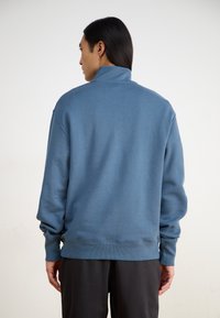 Felpa blu con colletto alto, polsini e orlo a costine, texture morbida e vestibilità ampia. Indossata con pantaloni neri, presenta un design semplice.