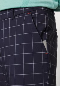 Mornarsko plave hlače s uzorkom mreže u bijeloj i svijetlo sivoj boji. Značajke uključuju bočne džepove, narančasti akcent i glatku, teksturiranu završnu obradu.