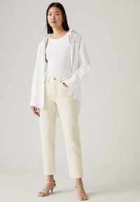 Witte oversized blouse gedragen over een witte tanktop, gecombineerd met crèmekleurige high-waisted boyfriend jeans en nude haksandalen. Soepele texturen, relaxte pasvorm.