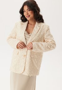 Beige texturerad blazer med fluffigt tyg, två främre fickor och en enkel knappstängning. Bärs över en ljusfärgad topp.