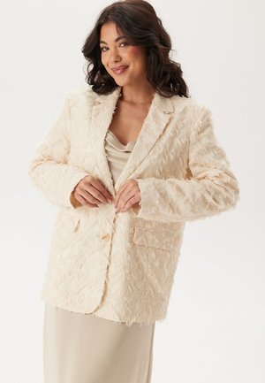 Beige tekstureret blazer med fluffy stof, to frontlommer og en enkelt knaplukning. Båret over en lysfarvet top.