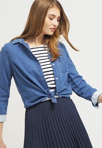 Denim skjorte med knaplukning, knyttet i taljen over en stribet t-shirt, parret med en let, plisseret mørkeblå nederdel. Blød tekstur, afslappet design.
